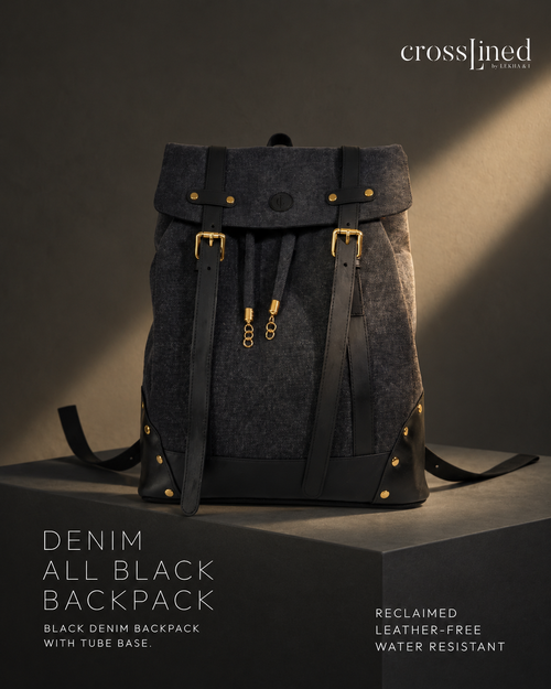 A Cross Lined backpack in dark denim and black tyre tube, displayed on a dark pedestal with dramatic side lighting. The backpack features a flap top with brass-tone buckles, drawstring details, and gold-tone rivets.