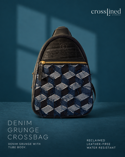 A Cross Lined crossbody bag made from reclaimed denim grunge, featuring a geometric 3D cube pattern in shades of blue and gray, with a black tyre tube top and gold zipper. The bag is displayed against a dark teal-blue background with soft window shadows.