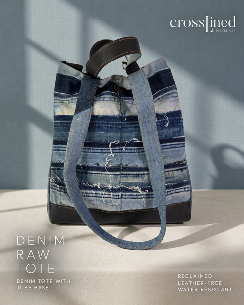 A denim raw tote bag with a distressed blue and white striped pattern, featuring dark tyre tube in-seam handles and base, displayed on a light beige surface against a textured blue-gray wall with dramatic shadows. The brand name 'crosslined by lekha & i' is visible in the top right corner.