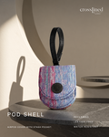 A small crosslined brand POD SHELL airpod cover with a stash pocket, featuring a textured blue, pink, and purple patterned fabric body with a black leather handle and snap closure, displayed on a dark stone pedestal with warm shadow lighting.