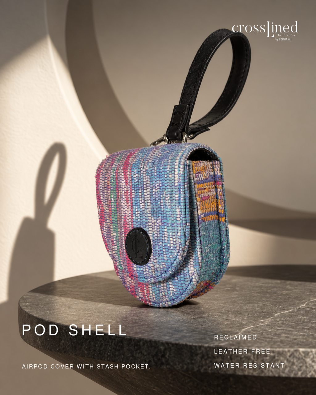 A small woven airpod cover pouch with a black wrist strap stands on a dark stone surface against a warm beige wall with strong sunlight and shadows. Text on the image reads 'crosslined', 'POD SHELL', and 'AIRPOD COVER WITH STASH POCKET.