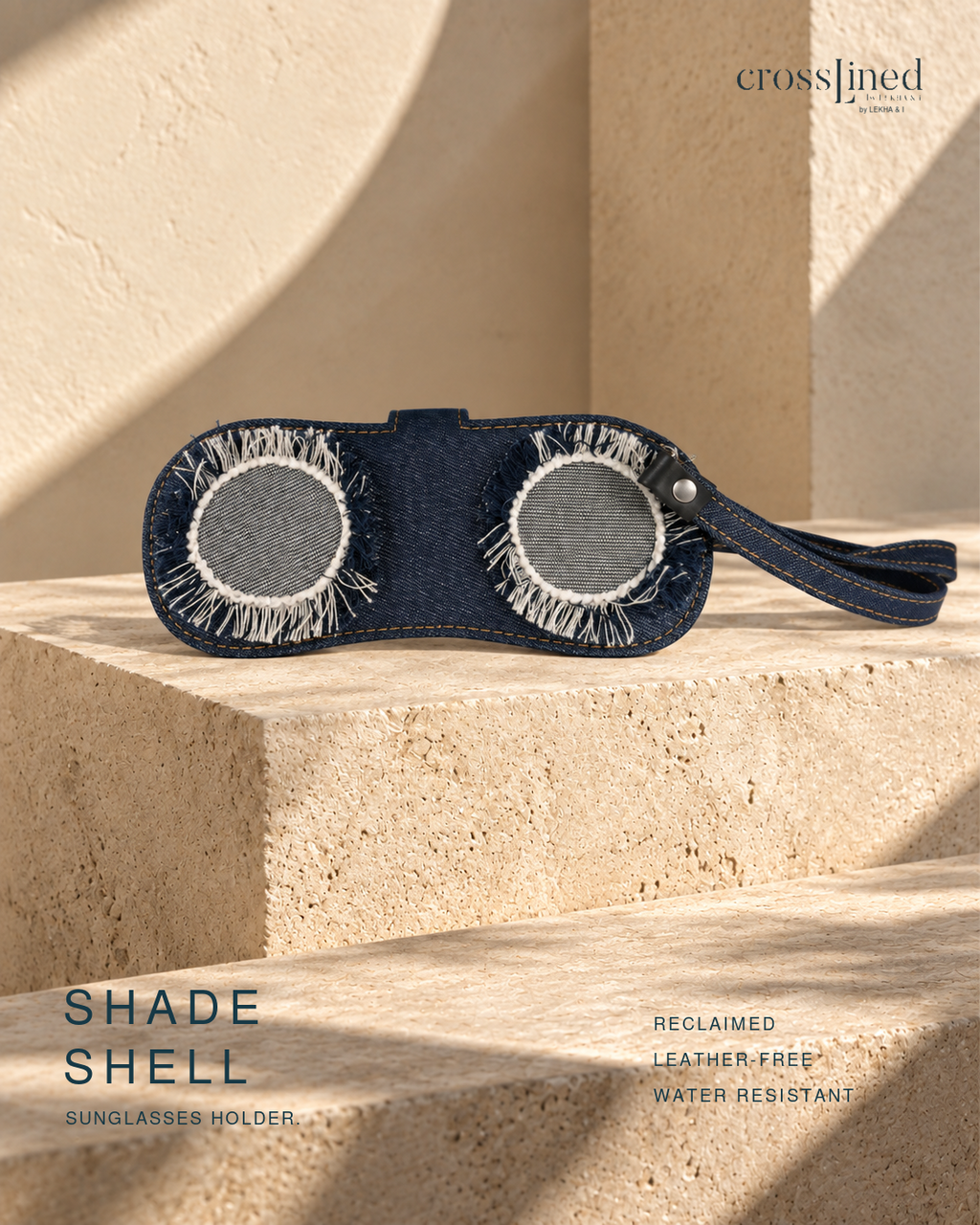 A dark blue denim sunglasses holder with two circular frayed fabric inserts and a wrist strap, displayed on a beige stone pedestal with architectural shadows. Text overlays read 'crossjined', 'SHADE SHELL', and 'SUNGLASSES HOLDER' with additional feature text on the right.