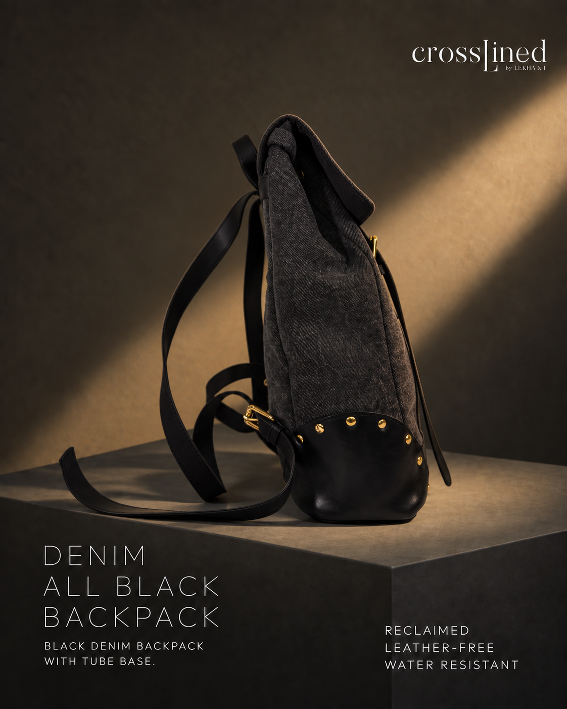A Crosslined reclaimed all-black denim backpack with a tube base, styled on a dark pedestal under dramatic warm lighting. The backpack features a dark gray textured denim body, a black leather base, and gold-tone rivets and hardware.