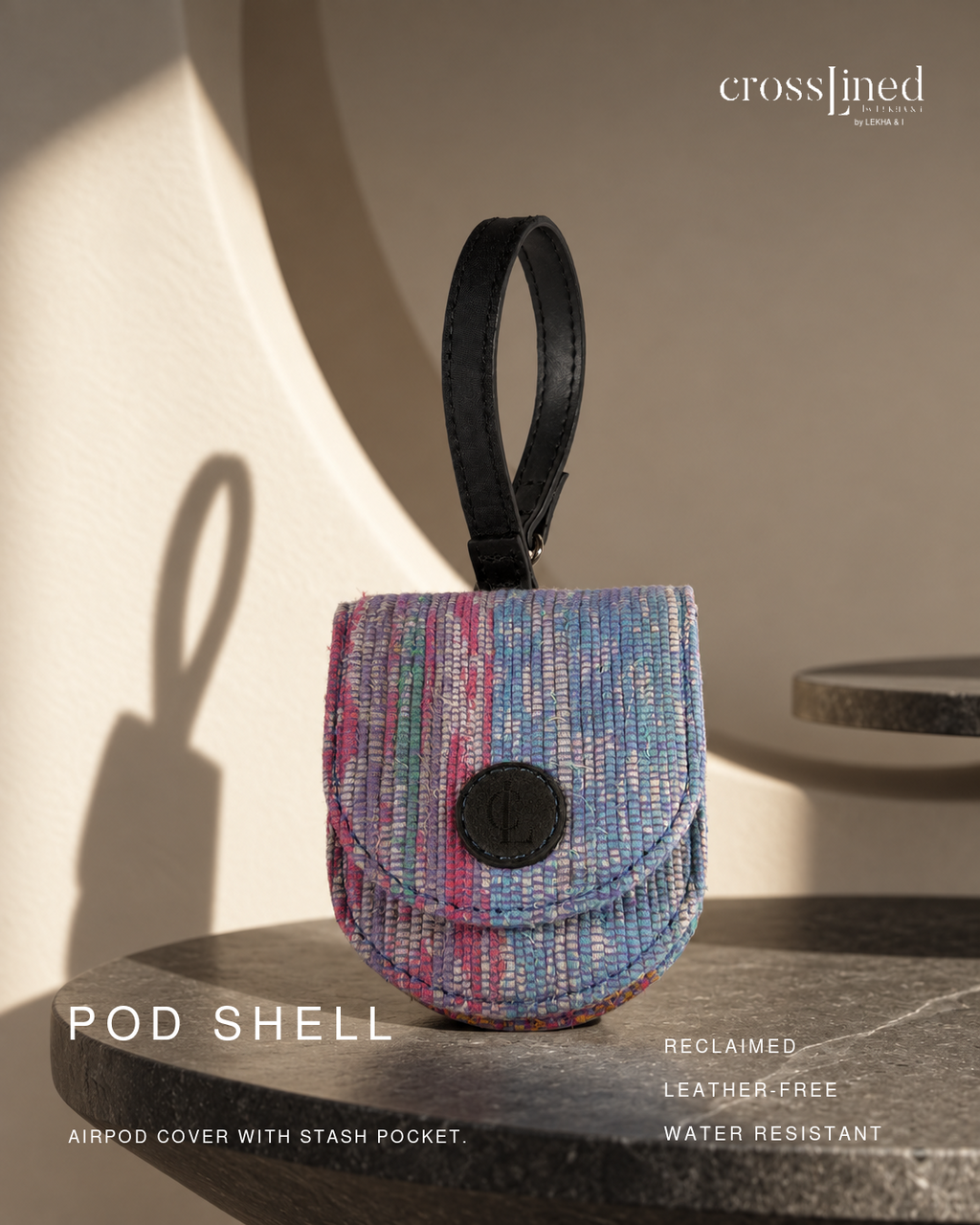 A small crosslined POD SHELL airpod cover with a stash pocket, featuring a textured multicolor woven pattern in blue, pink, and purple with a black leather loop and black snap closure. The product is displayed on a dark stone surface with dramatic warm sunlight and shadows against a beige background.