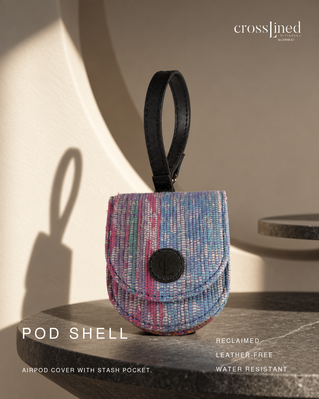 A small crosslined POD SHELL airpod cover with a stash pocket, featuring a textured multicolor woven pattern in blue, pink, and purple with a black leather loop and black snap closure. The product is displayed on a dark stone surface with dramatic warm sunlight and shadows against a beige background.