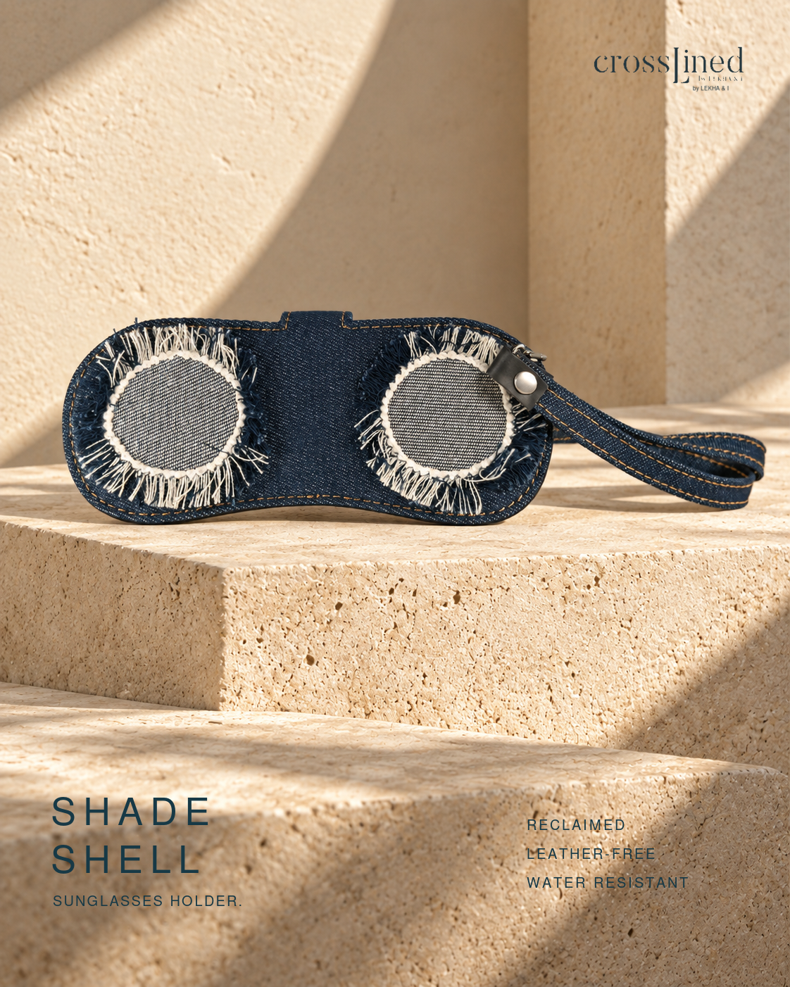 A navy blue denim-style sunglasses holder with two circular cutouts and fringe detailing, shown resting on beige stone steps in warm sunlight. The product is branded with 'crossjined' and labeled 'SHADE SHELL'.