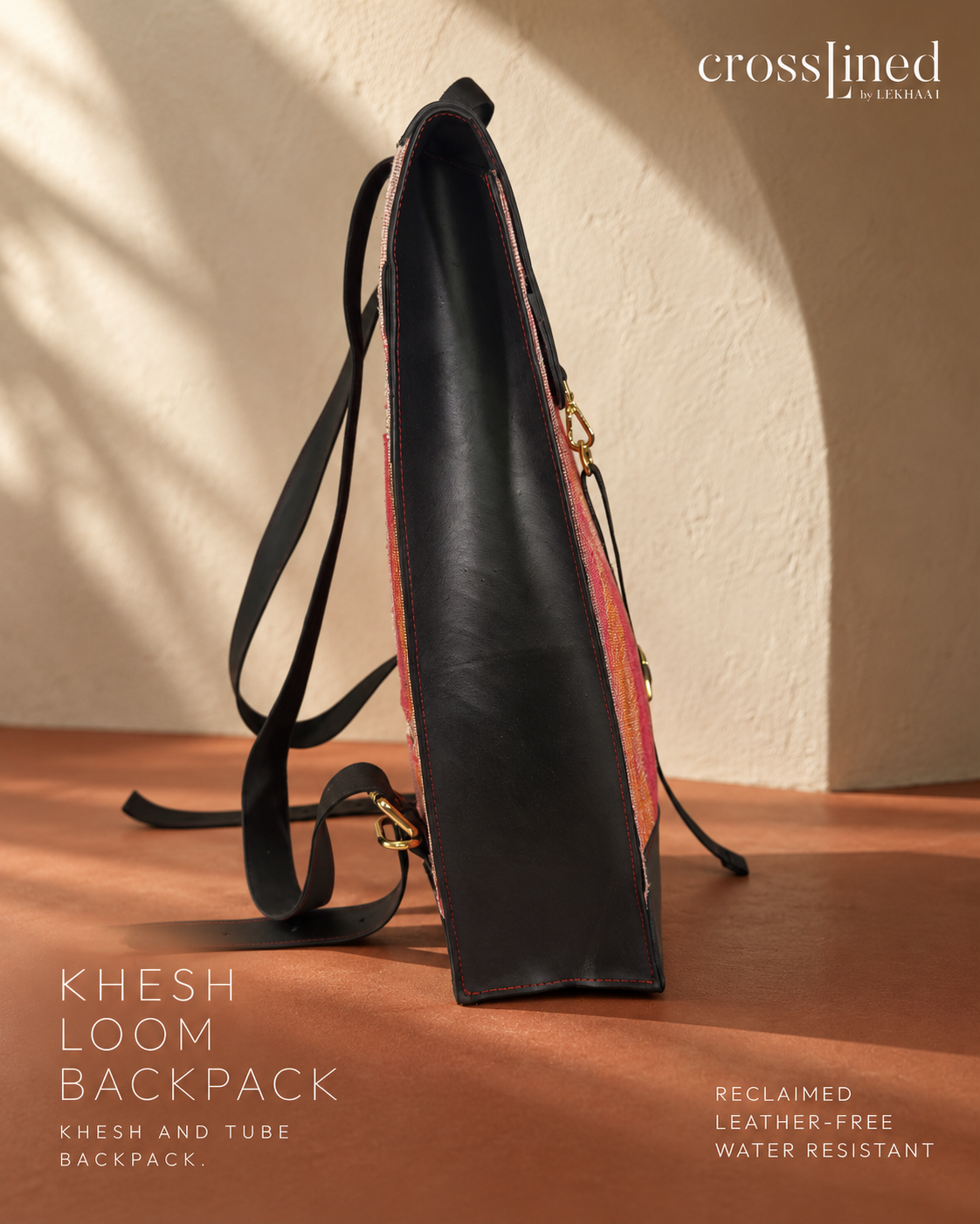 A CrossLined backpack shown standing upright on a wooden surface against a textured beige wall, with warm sunlight casting shadows. The backpack has a black leather-like side panel made from reclaimed tyre tube, a pinkish-red patterned interior, and gold-tone hardware.