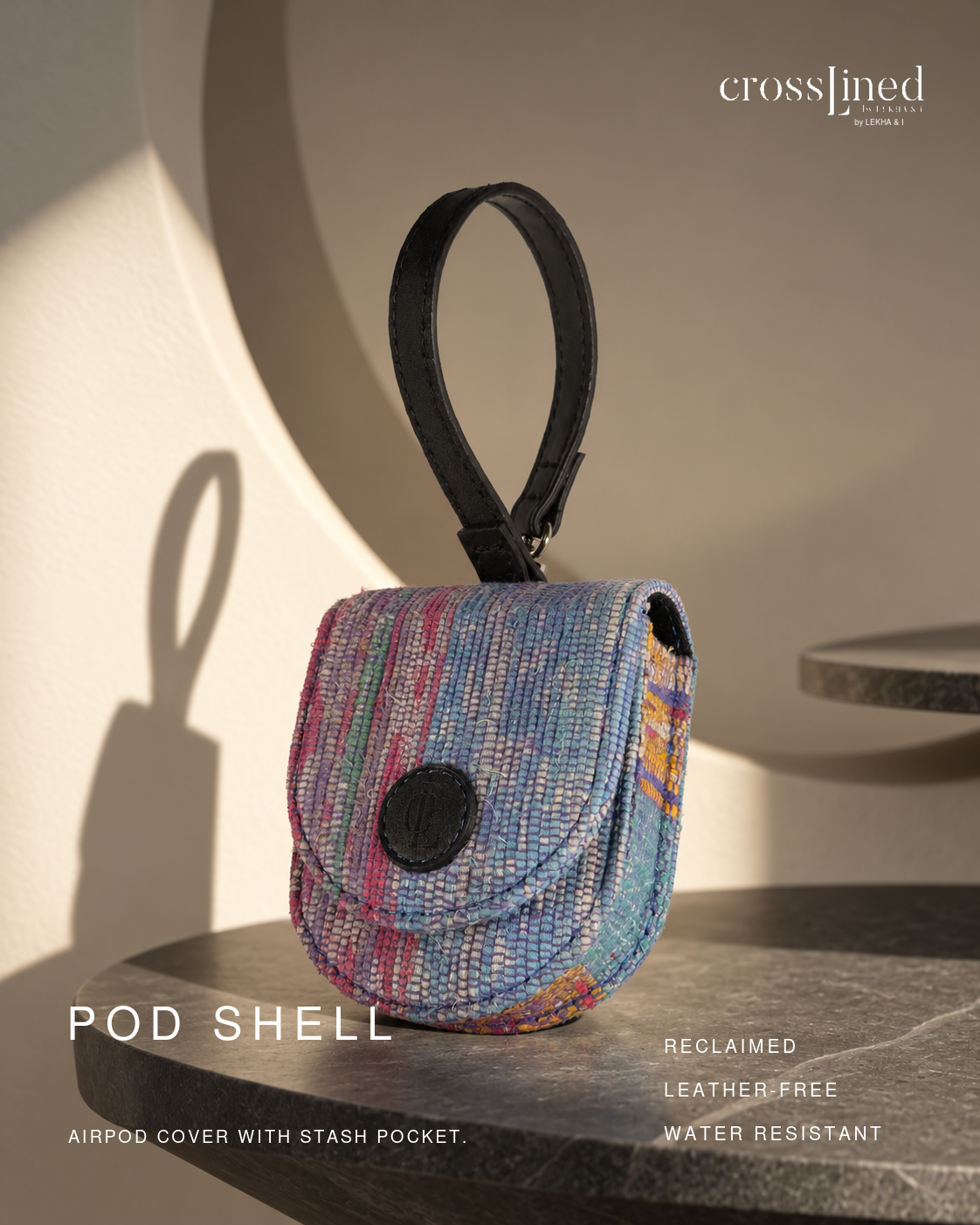 A small woven pouch cover with a black loop handle, shown on a stone surface in a styled interior setting. The pouch has a multicolor woven pattern with blue, pink, purple, and orange tones, plus a black circular snap closure.