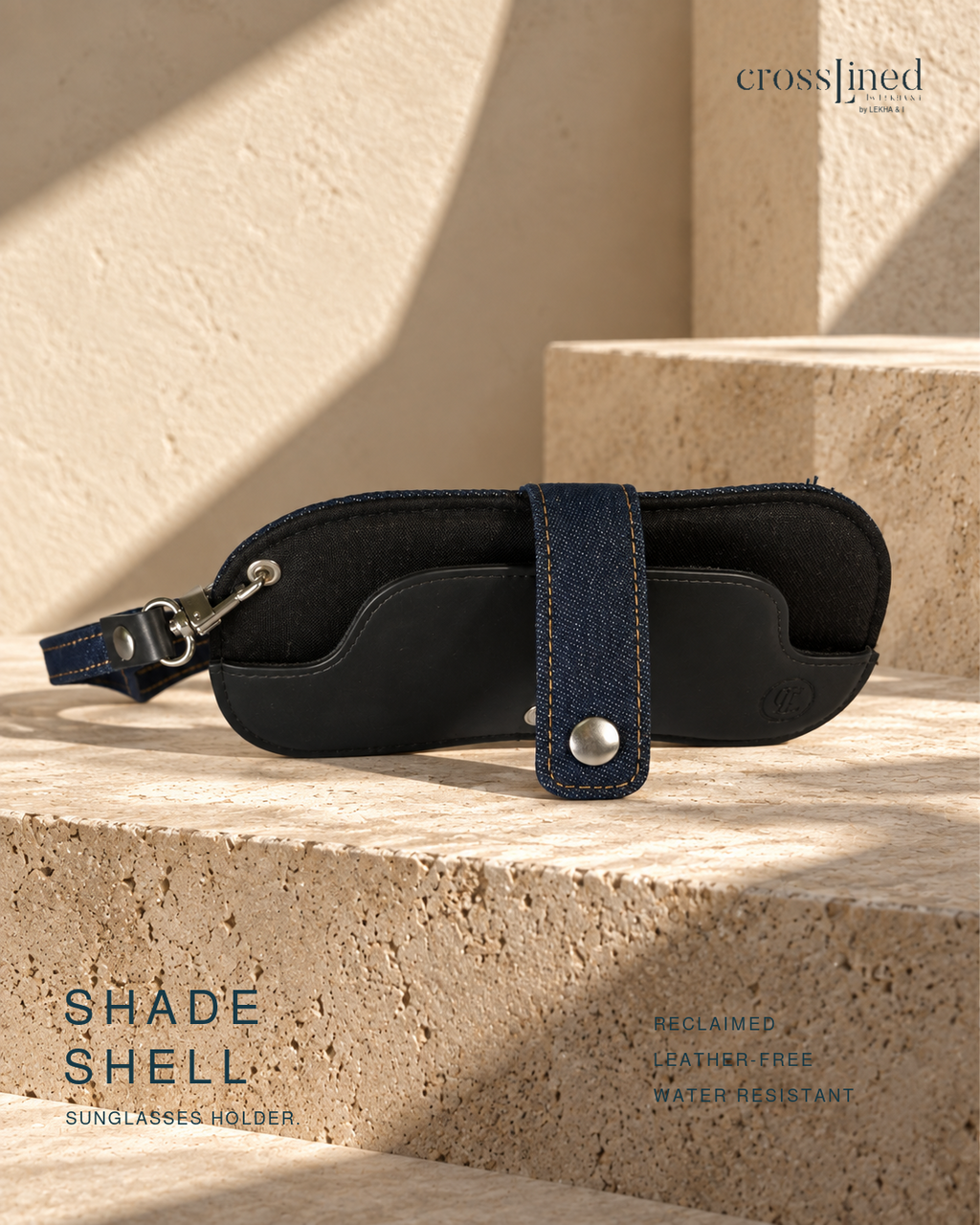 A black and navy blue sunglasses holder case with a dark denim strap and silver snap button, resting on a textured beige stone block. The brand name 'cross[jined' is visible in the top right, and 'SHADE SHELL' is written on the bottom left.