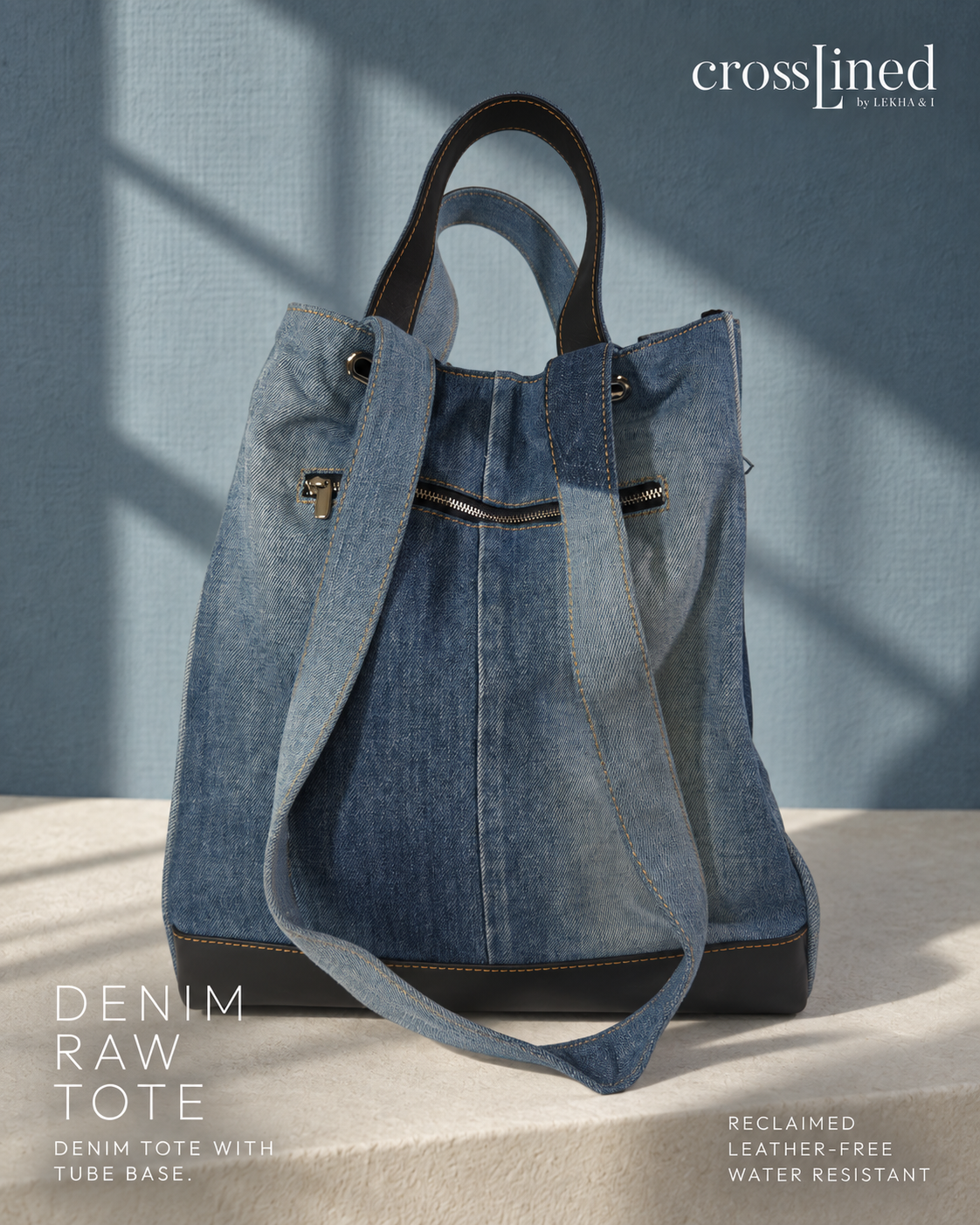 A Crosslined denim tote bag in a faded blue raw denim finish with black tyre tube inseam handles and trim, styled on a textured beige surface against a blue wall with soft window shadows.