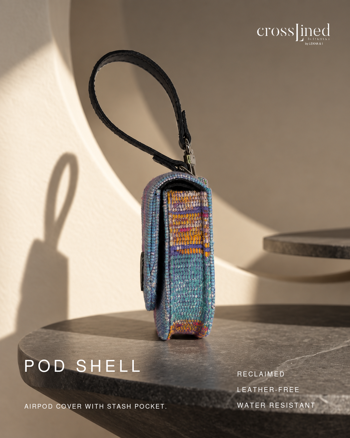 A small woven airpod cover pouch with a black wrist strap, shown standing on a dark stone surface in warm sunlight. The product has a multicolor woven pattern with blue, purple, orange, and beige tones.