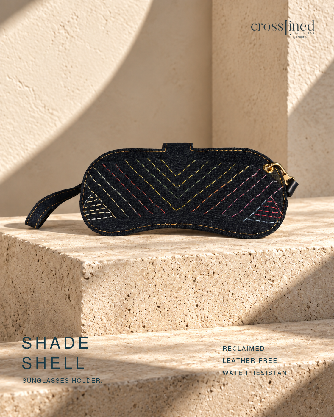 A dark navy sunglasses holder with colorful geometric embroidery and a gold hardware, displayed on a textured beige stone surface with dramatic shadows. The brand 'crosslined' is visible in the top right, and text at the bottom reads 'SHADE SHELL SUNGLASSES HOLDER' and 'RECLAIMED LEATHER-FREE WATER RESISTANT'.