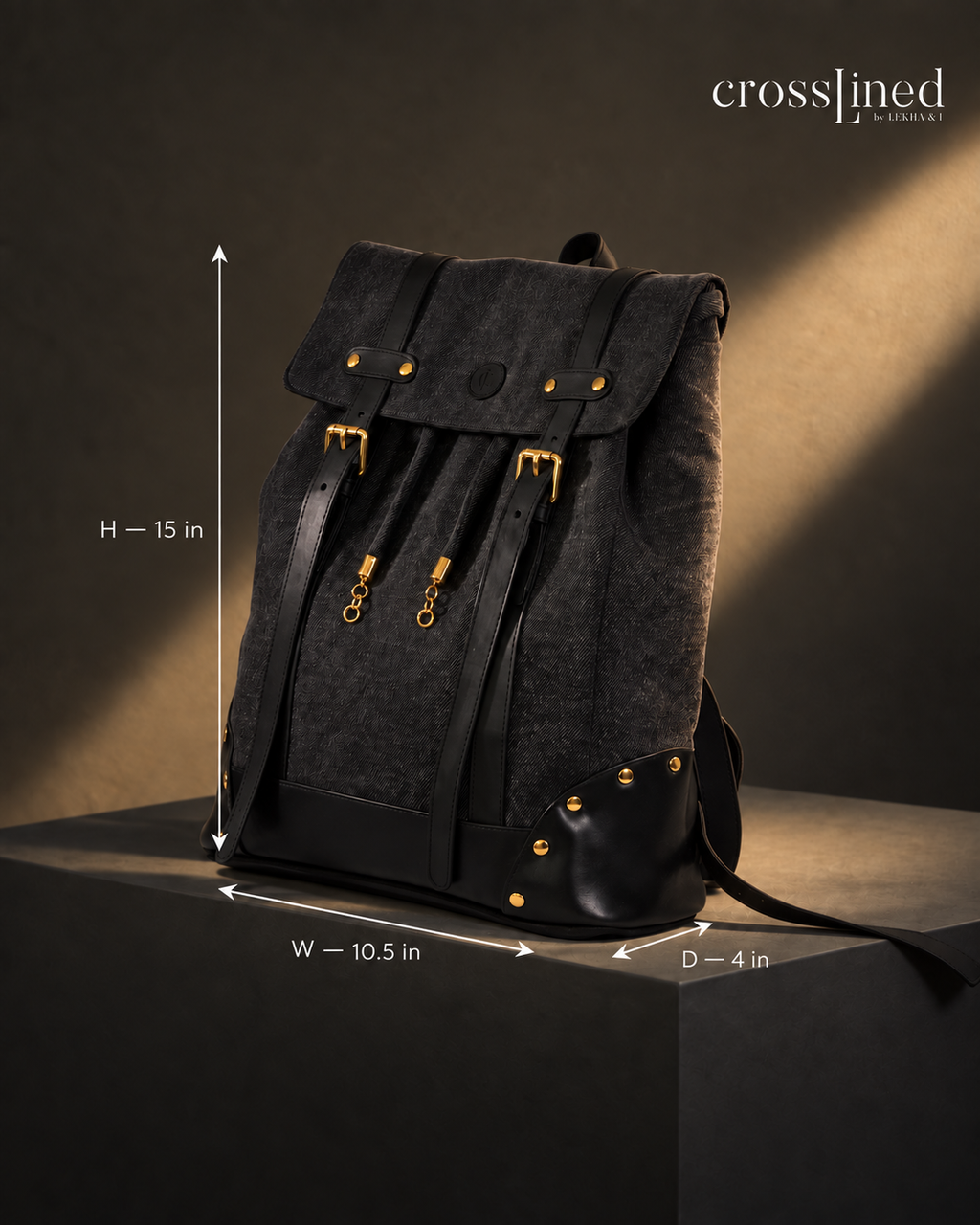 A dark charcoal black all denim backpack with black tyre tube straps and trim and gold-tone hardware, shown on a dark pedestal with dimension arrows and labels indicating height 15 in, width 10.5 in, and depth 4 in.