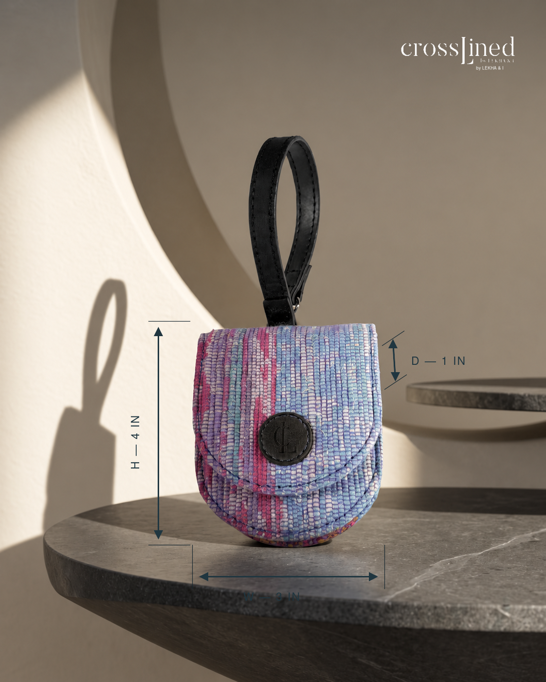 A small woven clutch or pouch with a black tyre tube handle and circular front clasp, shown on a stone shelf with dimension arrows and labels indicating height and width. The background is a warm beige wall with architectural shadows, and the Crosslined by Charlotte brand name appears in the top right.