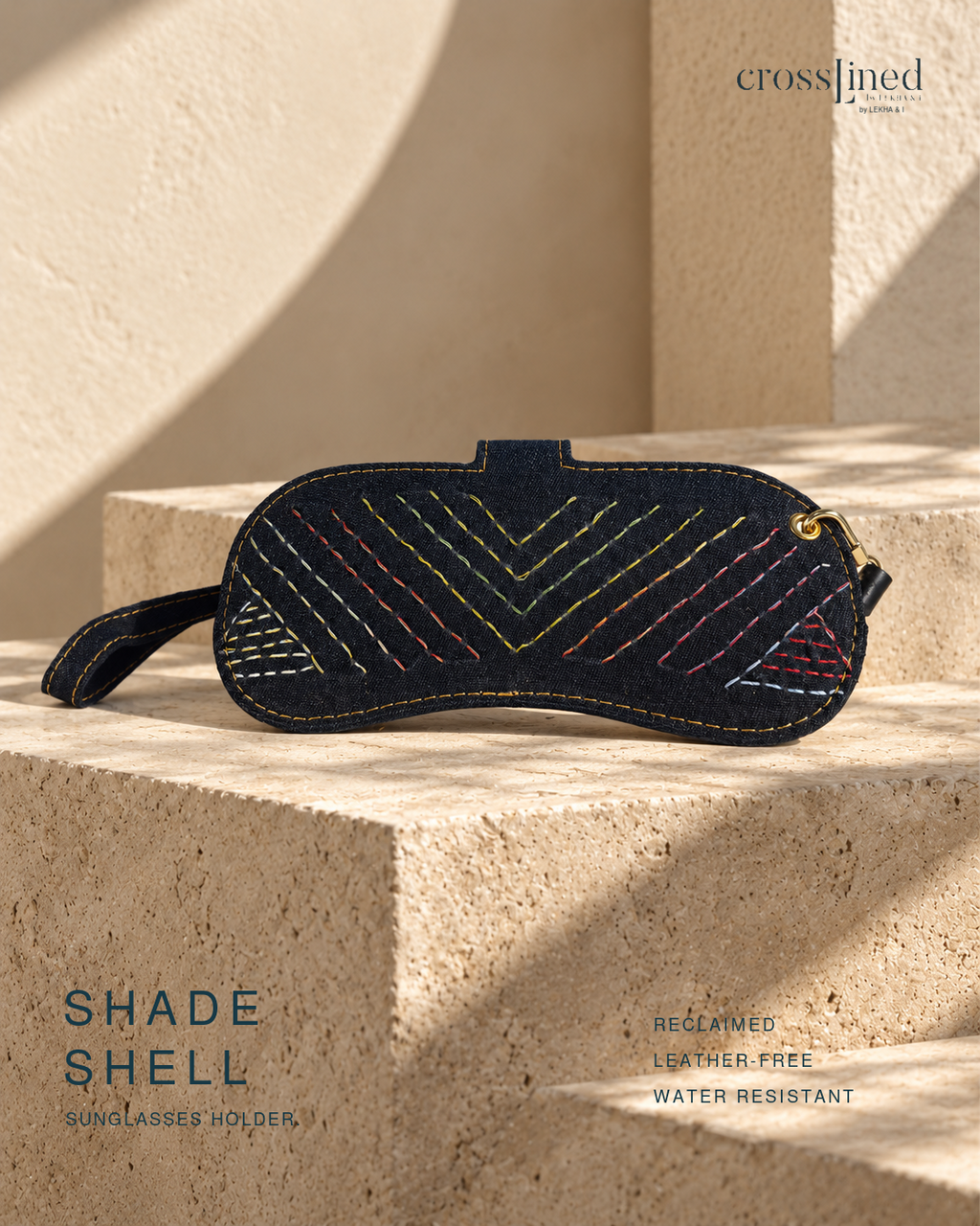 A reclaimed dark navy sunglasses holder with colorful embroidered geometric patterns, styled on a beige stone block under warm natural light. The brand name 'crossjined' is visible in the top right, and 'SHADE SHELL' is written on the bottom left.